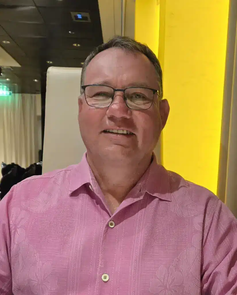 Man in glasses wearing a pink shirt sits indoors against a yellow-lit wall, facing the camera.