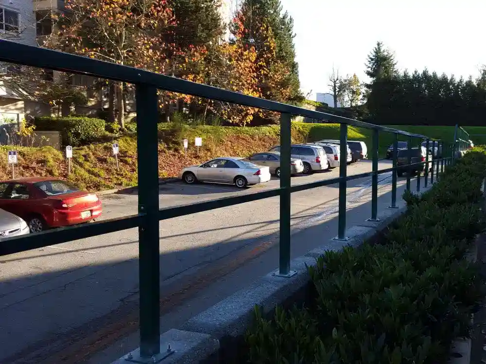 A row of parked cars lines a street next to a building, with outdoor railings and bushes in the