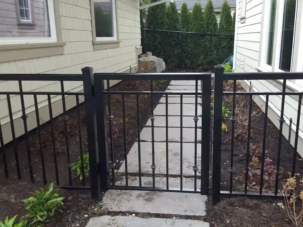 A black metal gate blocks a stone pathway between two beige houses, with sparse plants and soil on