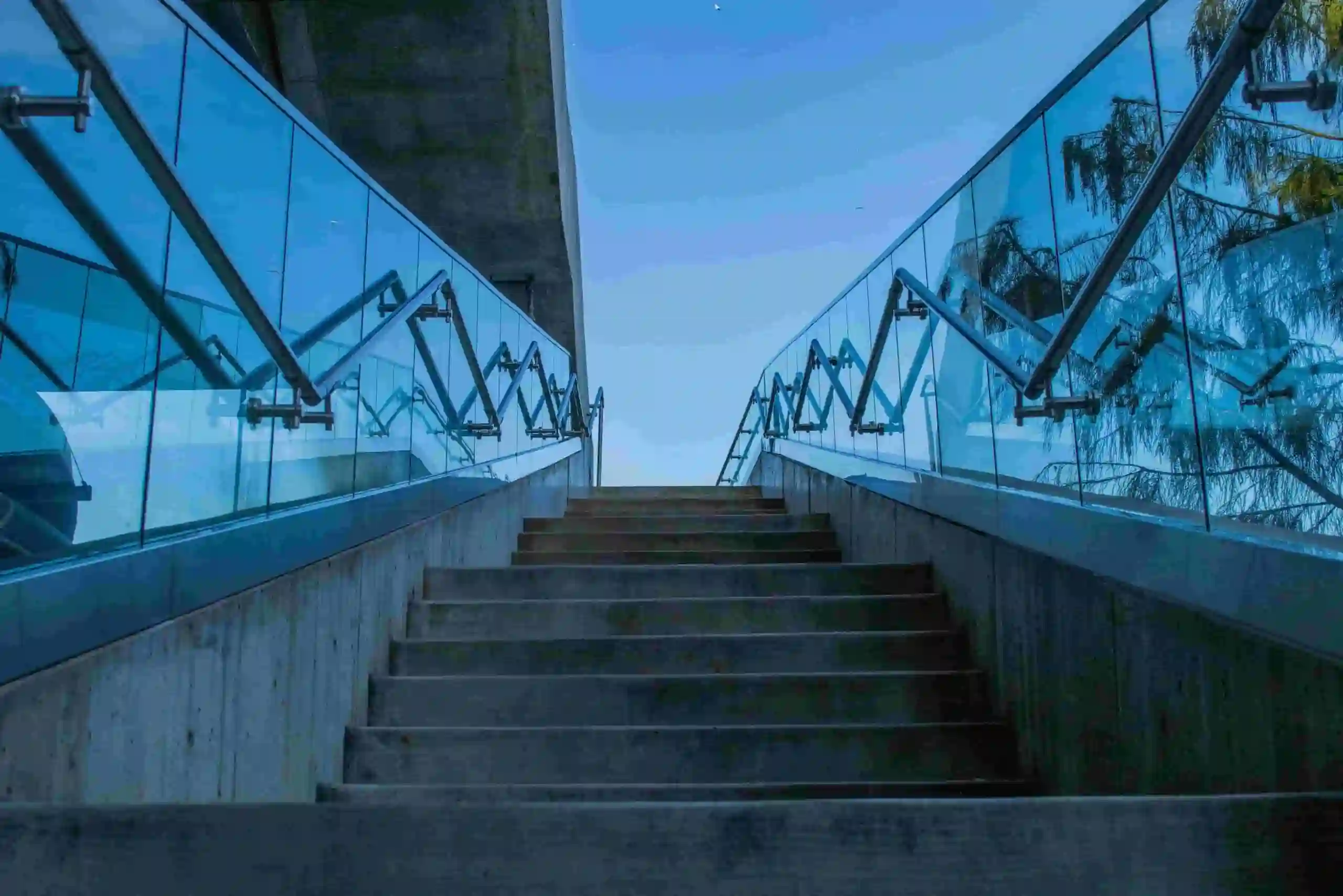 Concrete stairs lead upward, bordered by sleek glass railings with metal supports, under an open sky