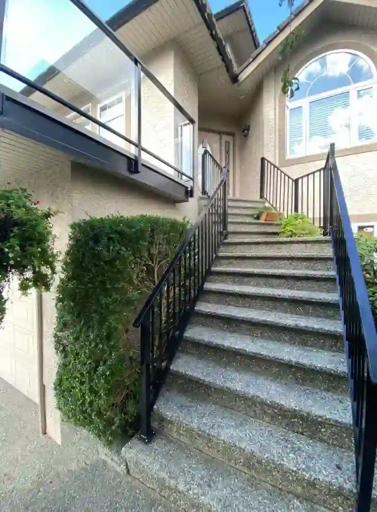 Concrete steps with a sleek metal railing lead up to the front door of a beige house, featuring a