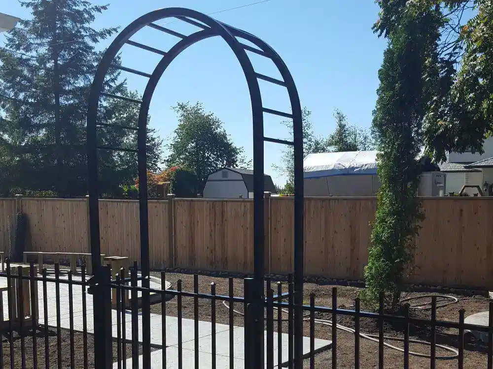 A black metal garden arch stands on a paved walkway in a fenced backyard with trees, a shed, and