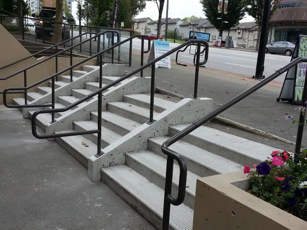 Concrete stairs with five steps and four black metal handrails, leading from a sidewalk up to a