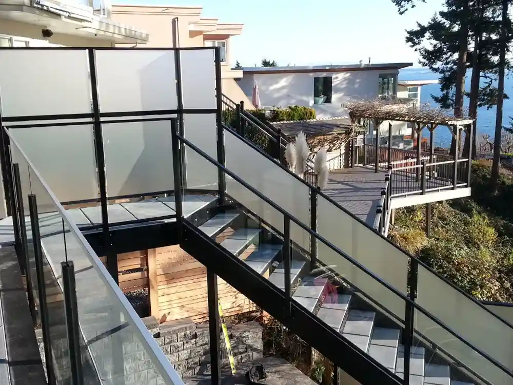 Glass-paneled stairway and balcony overlook a wooden deck with a pergola, surrounded by greenery