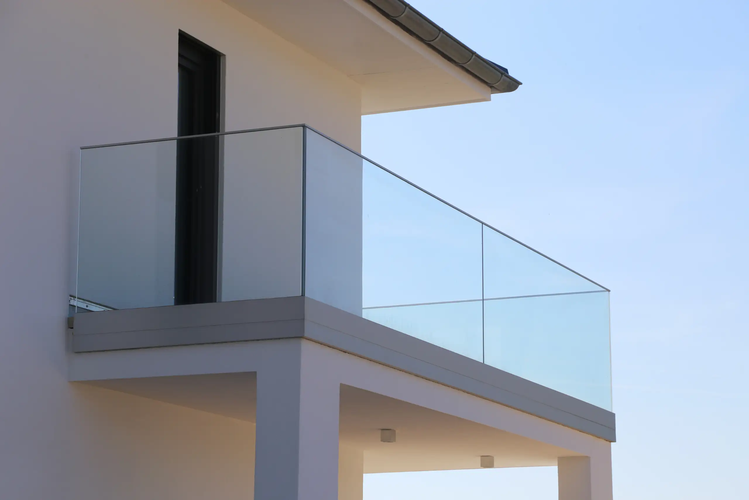 Modern house balcony with glass railings and white walls, viewed from an angle against a clear blue