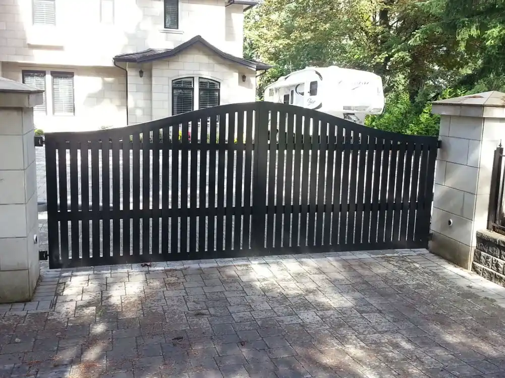 A black metal gate with vertical bars blocks the entrance to a stone driveway in front of a house
