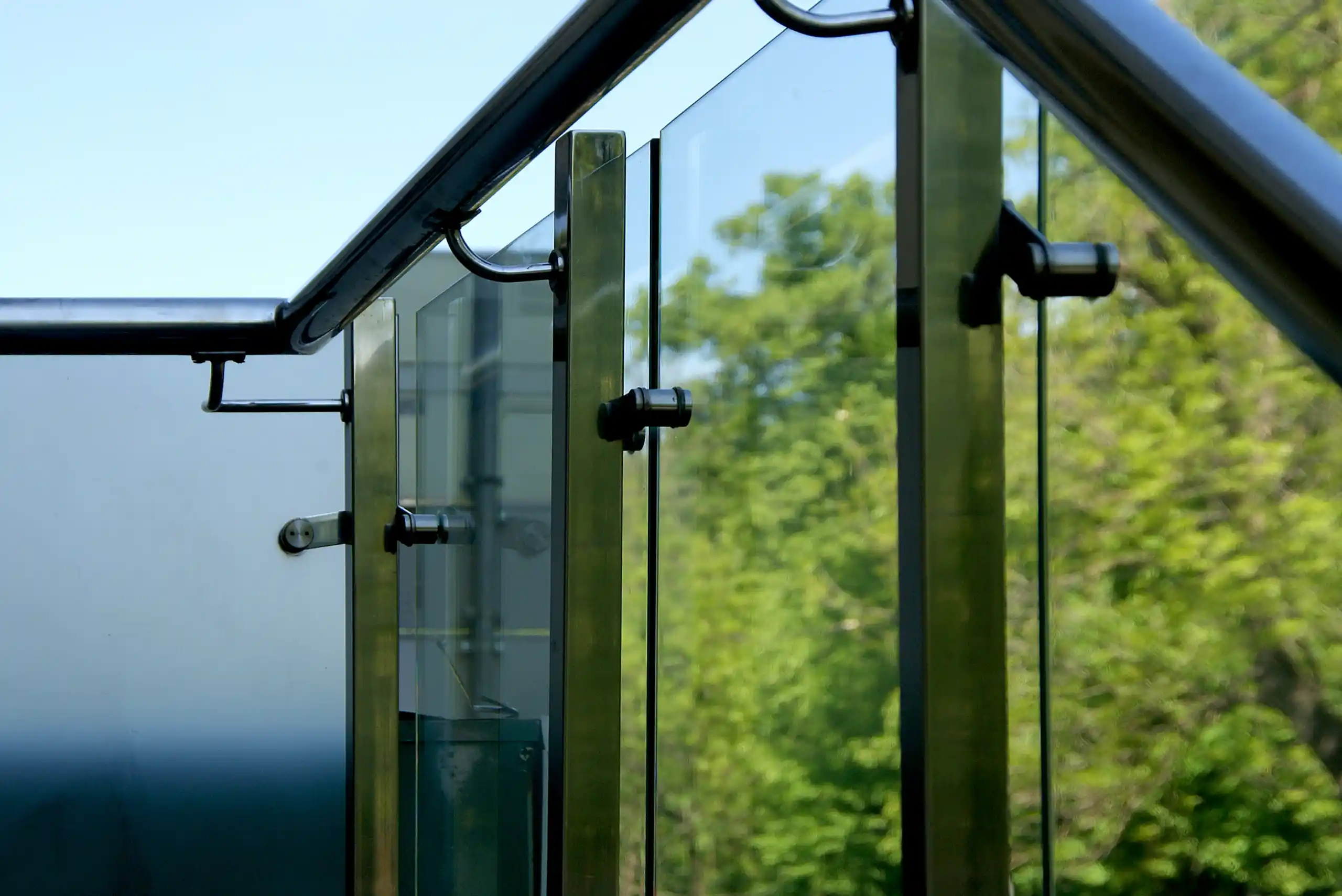 Close-up view of glass panels and metal supports on a structure with trees and blue sky visible in