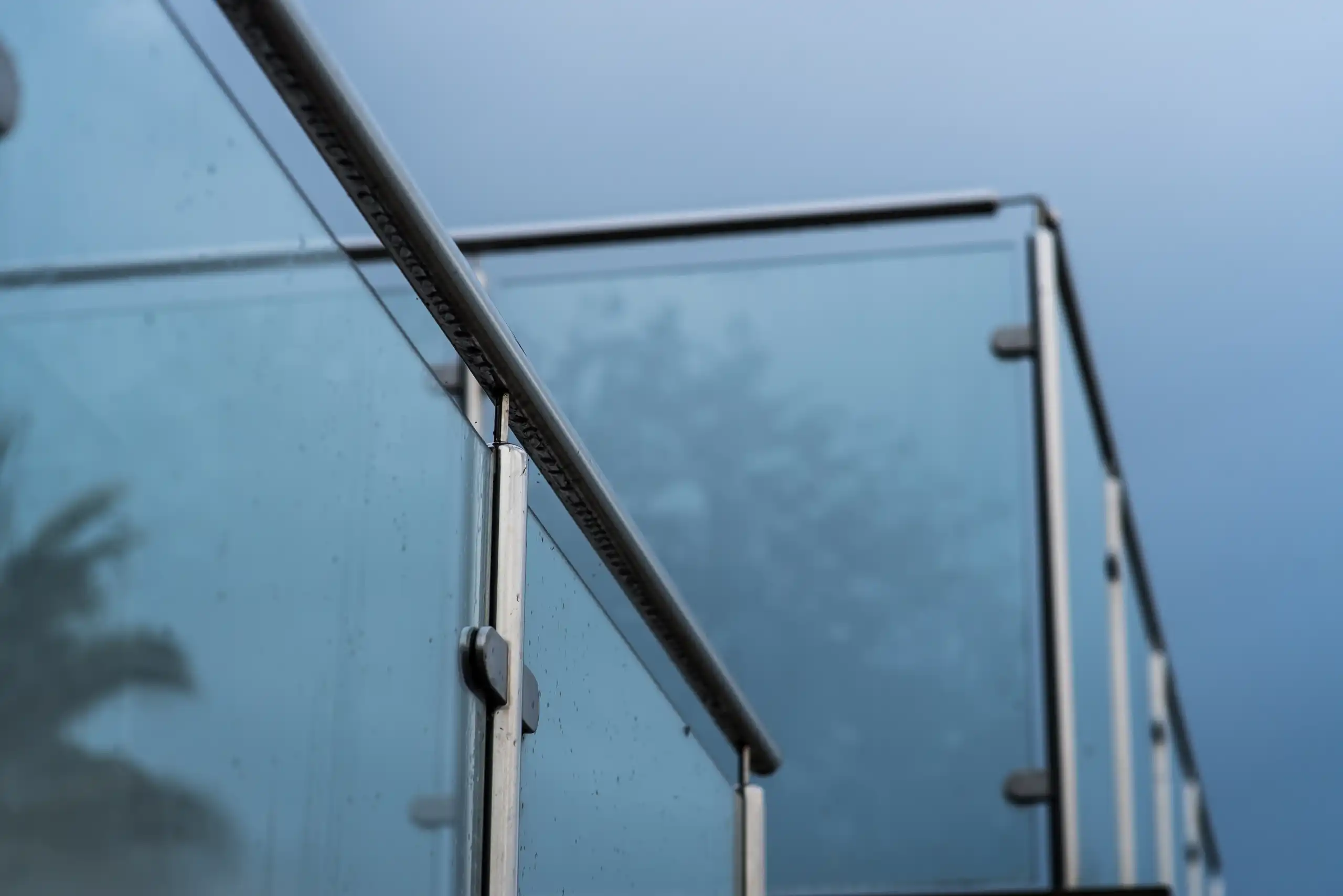 Close-up of a modern glass and metal balcony railing against a cloudy blue sky, with faint
