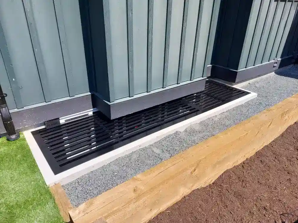 A black metal grate covers a drainage area next to a building with green siding, bordered by